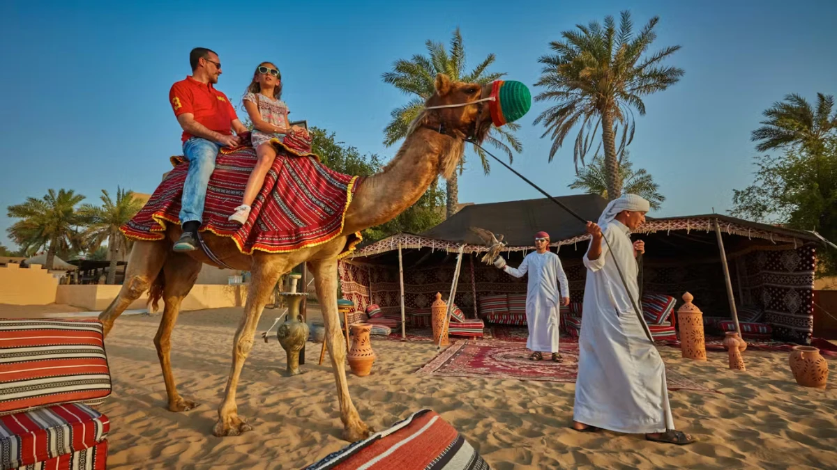 Couple riding a camel at a Dubai desert camp at sunset, guided by Bedouin staff with palm trees and tents behind.